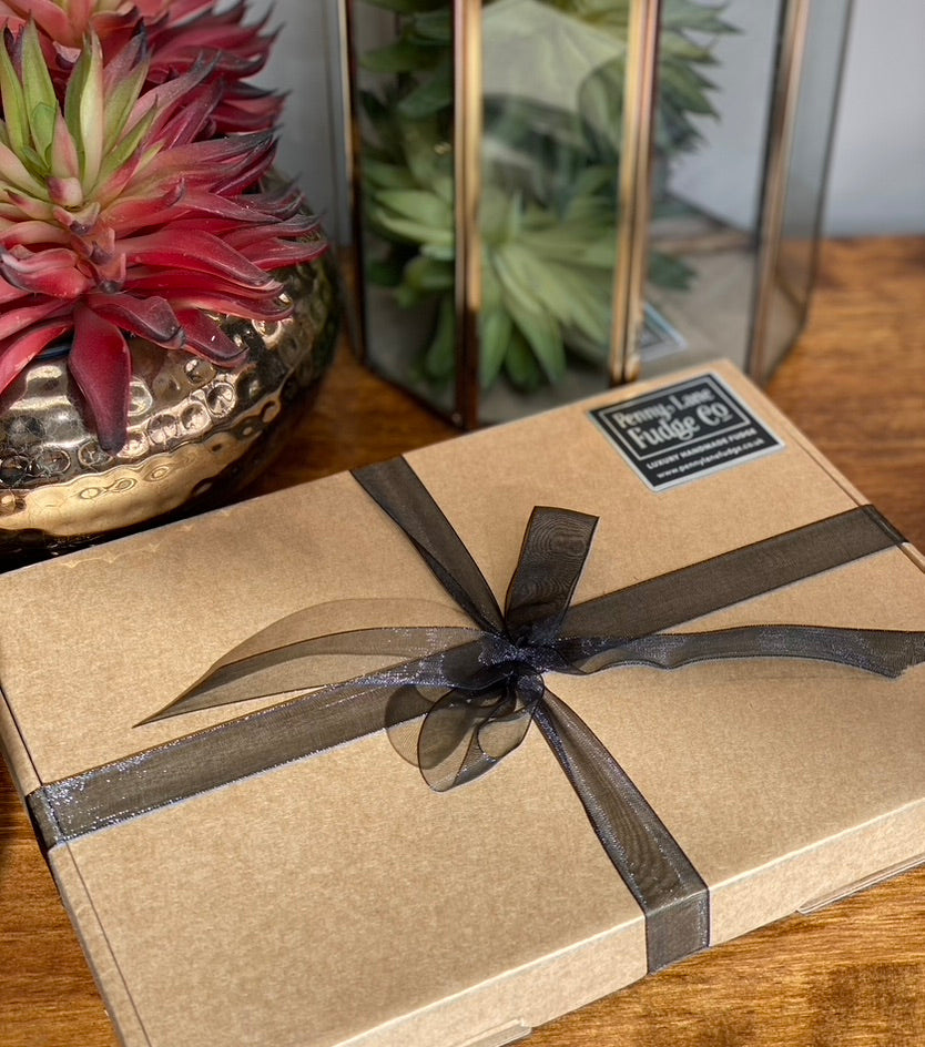 Gift box wrapped in brown paper with a black ribbon on a wooden surface, next to a plant.