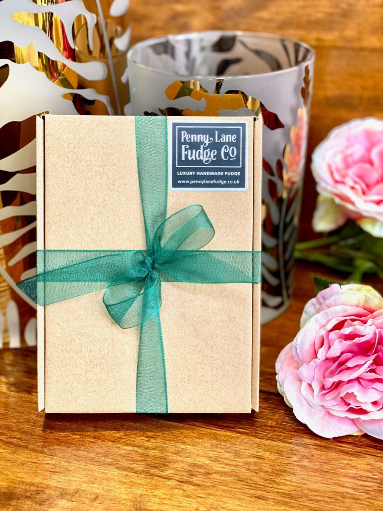 Gift box with a green ribbon and 'Penny Lane Fudge Co' label on a wooden surface with flowers.