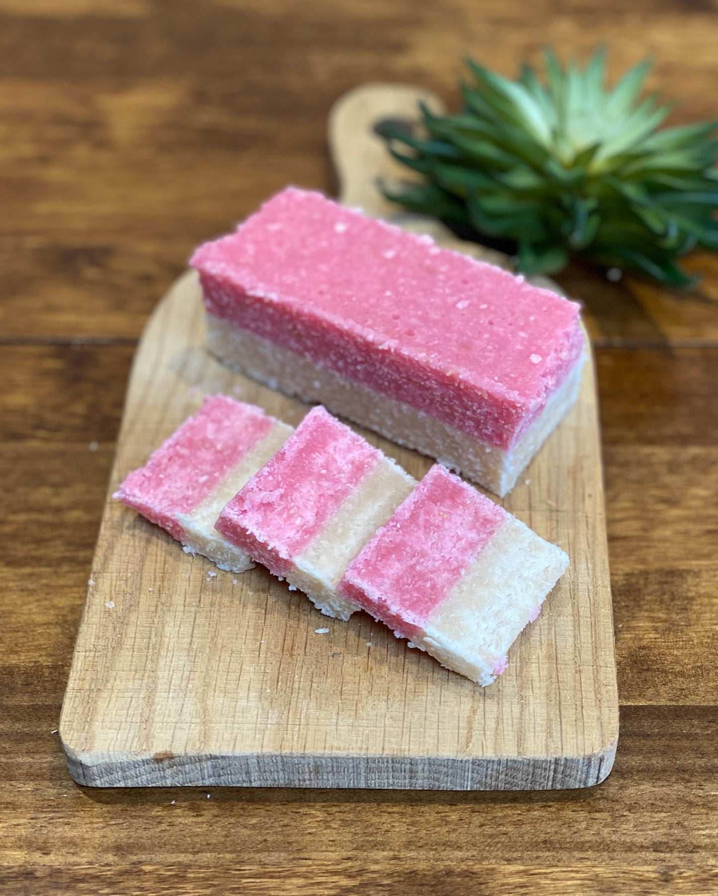Pink and white layered coconut ice bar on a wooden cutting board with a plant in the background.
