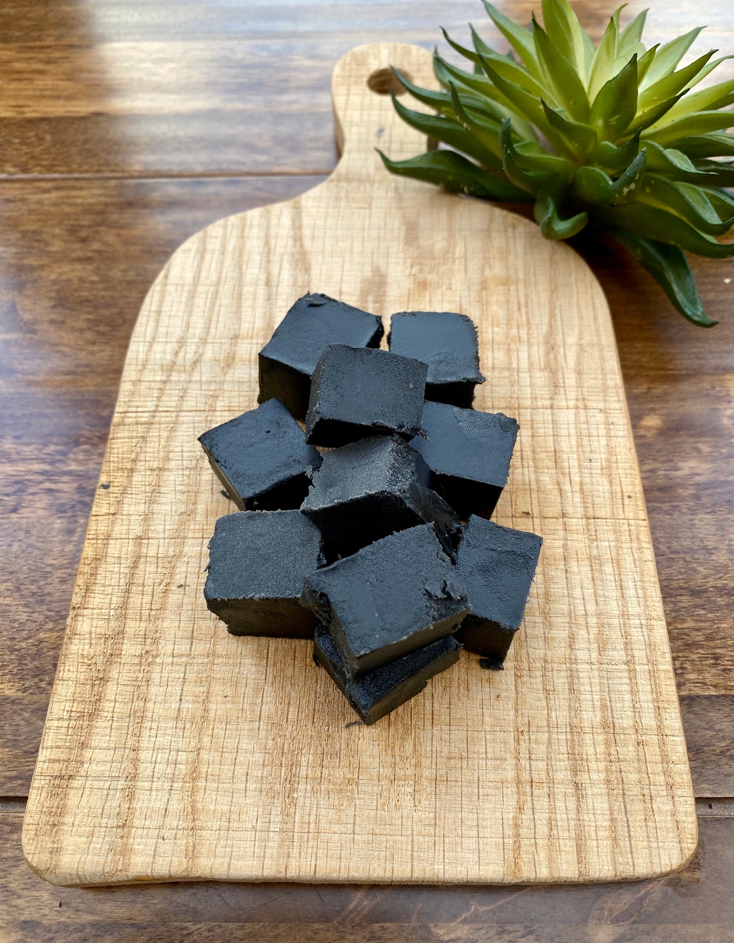 Black liquorice cubes on a wooden cutting board with a plant in the background